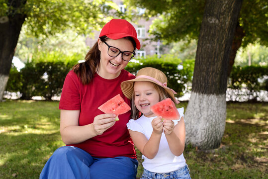 Smiling mother and daughter enjoying fresh watermelon together in a sunny park. Healthy summer snack, family bonding, outdoor leisure, and joyful childhood moments