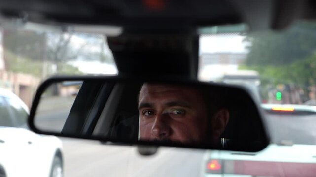 Eyes of male smiling driver reflecting in rearview mirror while riding motor auto. Young man looking concentrated to urban road during operating vehicle. Confident guy driving modern car at city