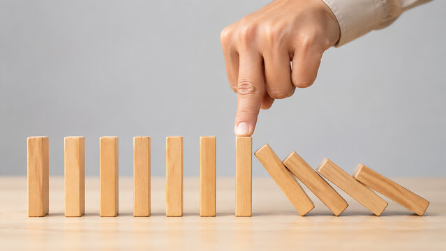 Hand stops the domino effect by pressing down on a light wooden block preventing the chain reaction of falling pieces in crisis management