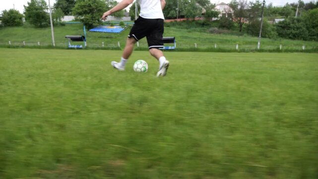 Legs of sportsman shows tricks with ball while running at green field. Male feet of professional footballer kicks ball during jogs at meadow on summer day. Young man training at stadium. Slow motion