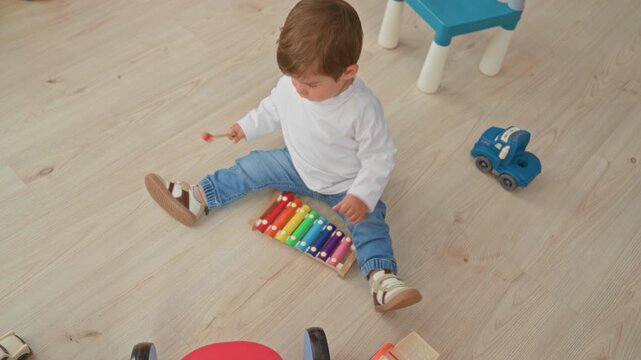 Boy sitting on wooden floor plays colorful xylophone with mallet near toy car and small chair in play area inside building; playful curiosity.