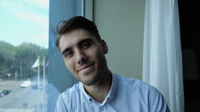 Portrait of happy tired businessman sitting near window in modern hotel room or office. Relaxed young man looks into camera with smile showing relief, confidence and success in professional lifestyle