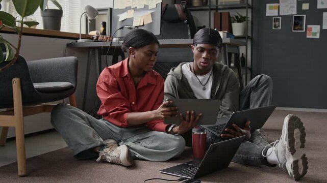 African American young programmers sitting on floor in creative office, discussing project on tablet and using laptops. Ideal for promoting diversity in modern IT workplace culture