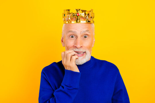 Nervous senior man wearing a golden crown and blue sweater biting his nails against a bright yellow background, anxious king portrait