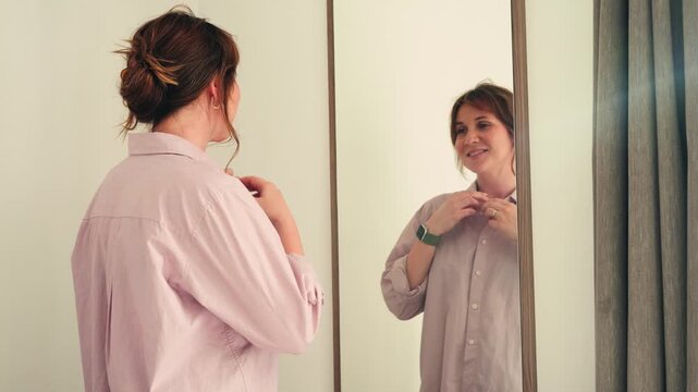 Young woman standing in front of a mirror at home and buttoning her shirt. Morning routine, self care, confidence and everyday lifestyle concept. High quality 4k footage