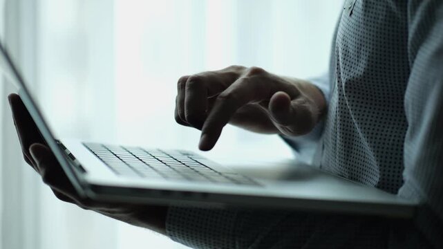 Male hand of young businessman typing text on laptop keyboard near window in modern office. Professional manager working on computer with natural light, showing productivity and communication