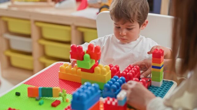 Boy stacking colorful plastic blocks with hands on a play table in a building classroom while a woman guides the tower; quiet concentration.