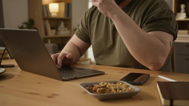 Cropped shot of Caucasian adult man with glucose monitor on arm typing on laptop and eating healthy nuts at home. Clip perfect for diabetes management and health technology marketing campaigns