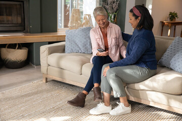 Obraz premium Mature female friends sitting on beige sofa in living room, leaning in showing smartphone