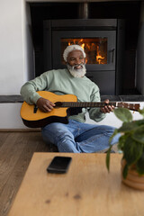 Obraz premium Senior African American man sitting on floor playing guitar by wood stove, smartphone on table