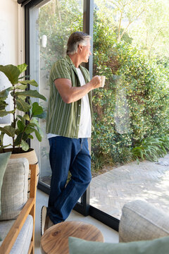 Senior man standing by sliding glass door holding white coffee mug and gazing at sunlit garden
