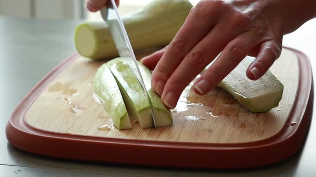 Close-up of a woman's hands using a sharp chef's knife to slice fresh green zucchini on a wooden cutting board in a bright kitchen