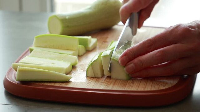 Close-up of a woman's hands using a sharp chef's knife to slice fresh green zucchini on a wooden cutting board in a bright kitchen