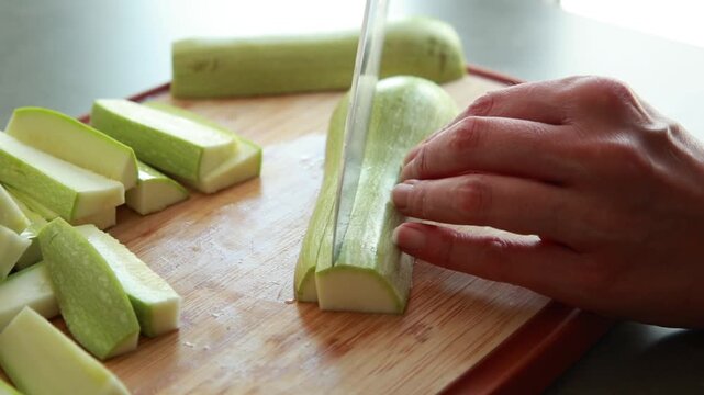 Close-up of a woman's hands using a sharp chef's knife to slice fresh green zucchini on a wooden cutting board in a bright kitchen