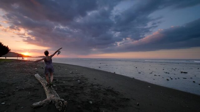 Slow motion: woman balancing on driftwood along Pemuteran Beach at sunset, Bali, Indonesia. Dramatic storm clouds, dark volcanic sand, and ocean waves create a serene coastal scene