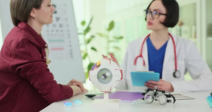 Human eye model and phoropter against doctor consulting patient during appointment in clinic. Lady ophthalmologist discusses test results with woman