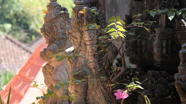 Close-up of a traditional Balinese guardian statue at a Hindu temple in Bali, Indonesia. Weathered stone carving adorned with a flower offering stands against lush tropical rainforest