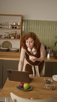 Vertical shot of pregnant Caucasian young adult woman eating breakfast while looking at laptop in kitchen. Clip ideal for maternal health, remote work, or nutrition campaigns