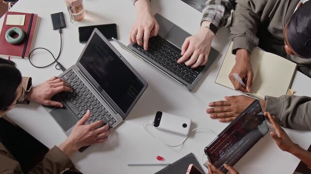 Top down shot of multiethnic programmers collaborating at desk, typing code on laptops and tablet while writing notes. Video suits tech startup promotions and software development marketing campaigns