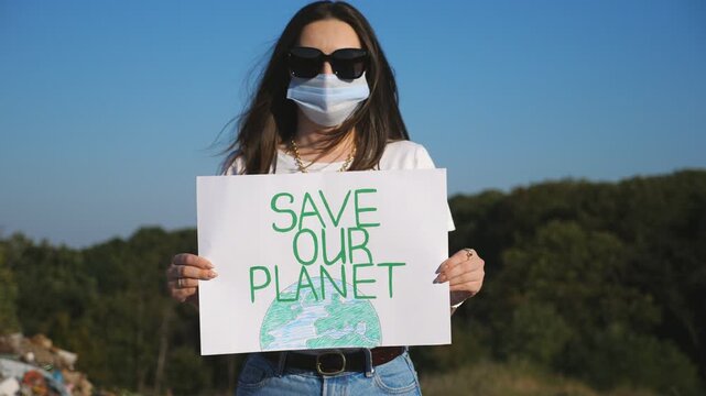 Portrait of young girl holding in hands poster of environmental movement for saving planet. Female eco volunteer in protective mask looking into camera and advocating for nature conservation. Close up