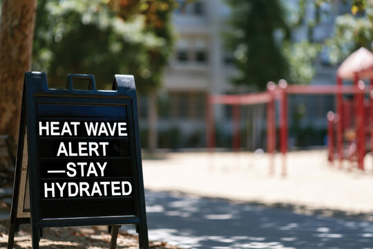 A public sign advising a heat wave alert and the importance of staying hydrated, set against a sunlit playground scene, promoting health awareness and community care during summer.