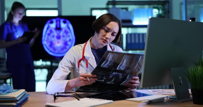 Focused doctor woman looks at patient spine x-ray images and works on computer in clinic. Physician analyzes injury danger with modern technology