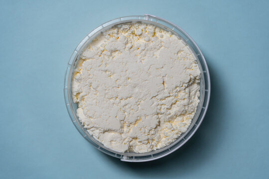 Fresh cottage cheese in a plastic bucket on blue background, closeup. Top view of organic white cottage cheese or curd in a round plastic container isolated on blue background
