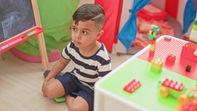 Boy sitting on floor with knee bent beside toy blocks and a chalkboard, gazing down in a playroom building; calm concentration.
