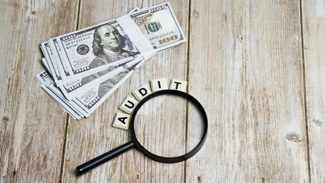 A magnifying glass placed over the word AUDIT spelled with letter tiles next to a stack of US hundred-dollar bills on a light wooden background.