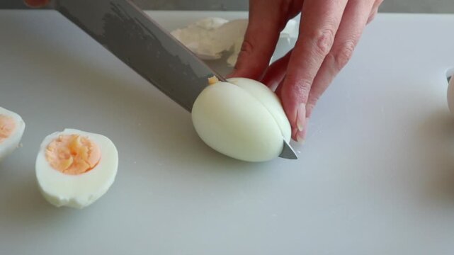 Close-up of a woman's hands cleanly slicing a hard-boiled egg in half with a sharp knife on a white cutting board