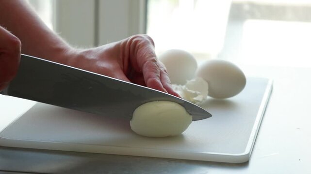 Close-up of a woman's hands cleanly slicing a hard-boiled egg in half with a sharp knife on a white cutting board