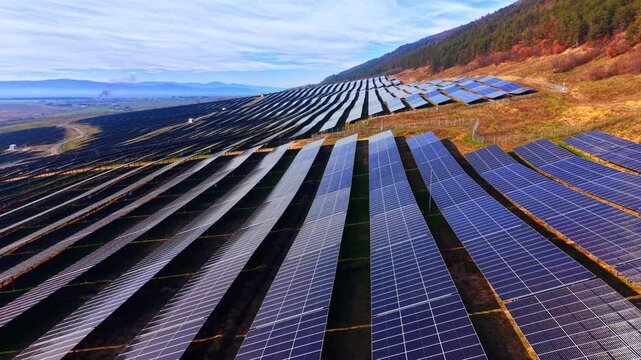 Converging solar panel rows facing mountain slope, Bulgaria. Low drone angle showing parallel blue arrays sweeping toward forested hillside with distant valley and overcast sky.