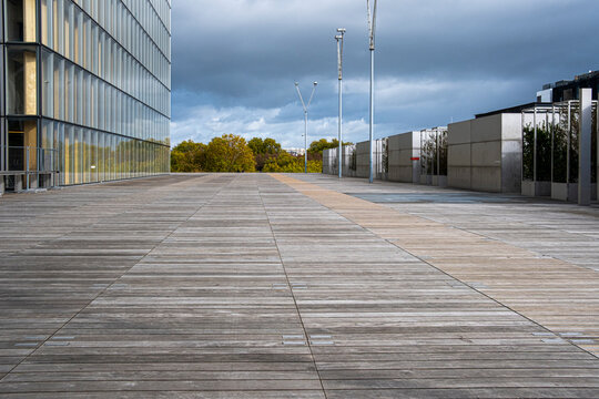 An empty wooden deck opens across an urban terrace, where architecture, storm clouds, glass facade, perspective, and plaza space stretch outward