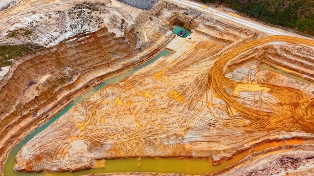 Drone view of layered sand quarry walls with teal water channel, Bulgaria. Oblique perspective showing orange and beige sediment layers, tire tracks, flooded trench and one vehicle on plateau.