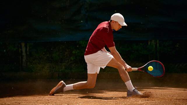Tennis player reaching low ball with backhand on dusty clay court. Concept of defensive tennis play, low ball control, mental focus and coaching lesson content.