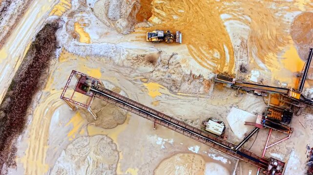 Overhead view of conveyor belt and wheel loader in sand quarry. Top-down drone shot of one yellow loader, rust-framed belt structure, beige and orange sand ground with tire patterns.