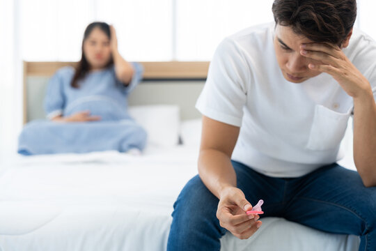 Man holding condom package while pregnant woman sits in background, representing family planning, responsibility, and lifestyle concept in cozy indoor bedroom environment with natural daylight