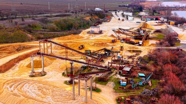 Aerial view of large sand and gravel plant with multiple conveyor lines. Wide drone perspective showing elevated belt frames, loaders, truck traffic, buildings and flat agricultural horizon.
