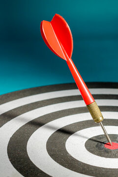 A vertical close-up shot of a single red dart firmly embedded in the red center bullseye of a dartboard against a blue background, representing ultimate success, perfect focus, and definitive victory.