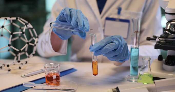 Female scientist pours reagent into test tube with yellow liquid in chemical lab. Technician in gloves researches color substance in laboratory