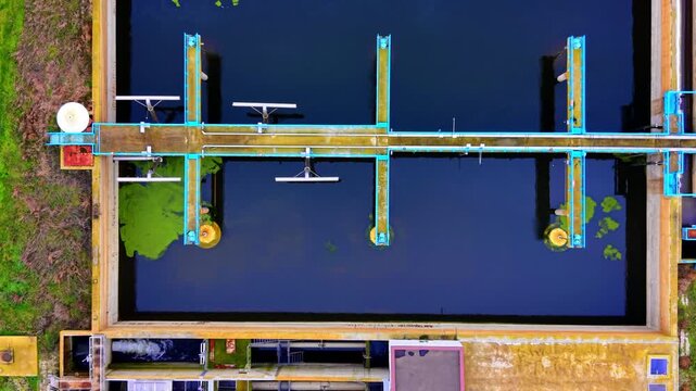 Overhead view of aeration basin with blue framework at sewage plant. Top-down drone shot of rectangular dark water tank with cyan pipe grid, surface aerators and yellow concrete walls.