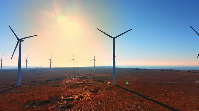 Sunrise light over wind farm on red coastal plain. Turbines framing sunburst, long shadows across russet earth, sea glimmering at horizon. Aerial view.