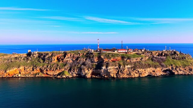 Lighthouse station on rocky headland at Cape Kaliakra, Bulgaria. Aerial view showing layered orange cliff face, white and red roofed buildings on green plateau top, teal sea below, blue sky.