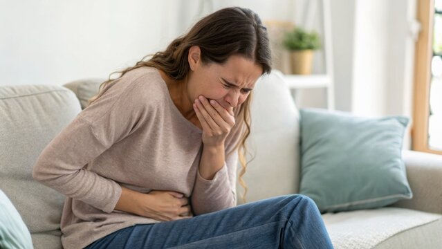 A woman sitting on a couch, clutching her stomach and covering her mouth, appears to be in pain or feeling nauseous in a bright living room.