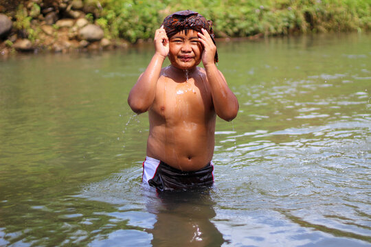 Indonesian boy smiling and playing in a shallow river wearing a traditional udeng headscarf.