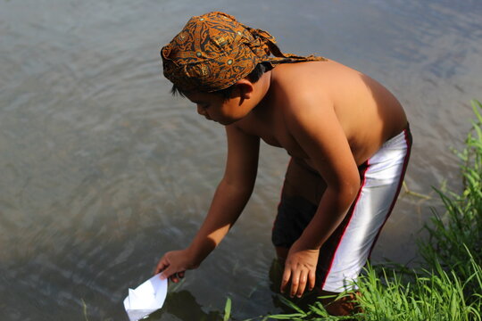 An Asian Indonesian boy wearing a traditional Javanese batik headtie plays with a white paper boat in a river, embracing culture and nature.