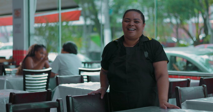 Latina waitress smiling warmly while standing in dining area, hand resting on chair in casual restaurant setting with guests in soft background
