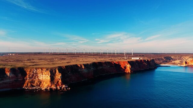 Red cliffs and wind turbines above calm Black Sea bay, Bulgaria. Sunlit orange cliff face center, flat blue bay below, wind turbine row on plateau top, wispy clouds in blue sky. Aerial view.