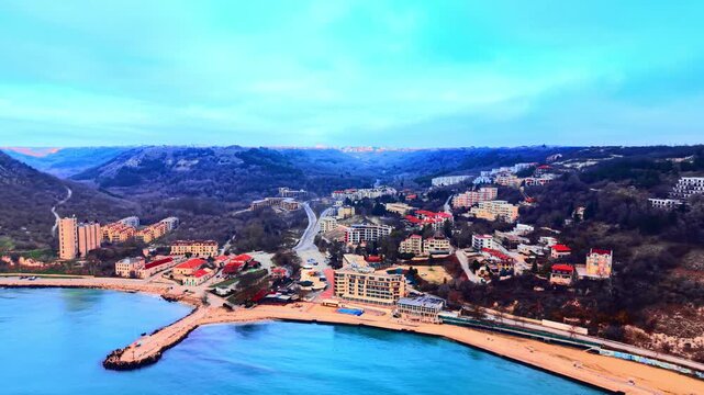Resort town and beach on hilly Black Sea coast, Bulgaria. Elevated oblique view, sandy shore and breakwater, hotels and apartments on forested valley slopes, city visible on distant ridge.