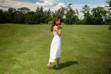 Obraz premium Girl in white dress holds strawberry and wants to eat it. Fragile Asian woman on picnic in park at dawn, summer vacation and outdoor recreation